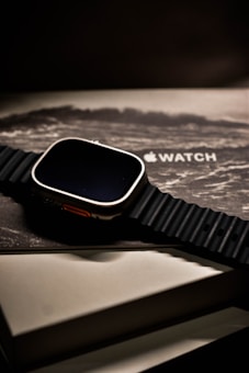 A sleek, modern smartwatch with a black ribbed band is placed on top of its packaging box, which shows an artistic wave design and the logo of the brand. The watch has a metallic casing and is presented at a slight angle, creating a sophisticated and luxurious appearance.
