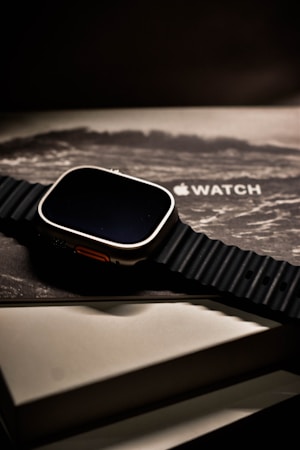 A sleek, modern smartwatch with a black ribbed band is placed on top of its packaging box, which shows an artistic wave design and the logo of the brand. The watch has a metallic casing and is presented at a slight angle, creating a sophisticated and luxurious appearance.