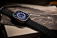 A smartwatch with a digital compass display rests on packaging, featuring a textured black strap. The background includes the word 'WATCH' and an illustration resembling a wave or abstract design.
