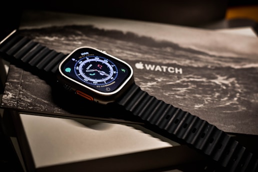 A smartwatch with a digital compass display rests on packaging, featuring a textured black strap. The background includes the word 'WATCH' and an illustration resembling a wave or abstract design.