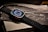 A smartwatch with a digital compass display rests on packaging, featuring a textured black strap. The background includes the word 'WATCH' and an illustration resembling a wave or abstract design.