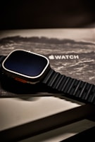 A smartwatch with a rugged black band rests atop its packaging. The watch face is dark, and the box beneath displays the product branding in a sleek, modern font. The overall aesthetic is minimalist and high-tech.