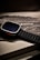 A smartwatch with a rugged black band rests atop its packaging. The watch face is dark, and the box beneath displays the product branding in a sleek, modern font. The overall aesthetic is minimalist and high-tech.