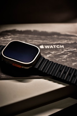 A smartwatch with a rugged black band rests atop its packaging. The watch face is dark, and the box beneath displays the product branding in a sleek, modern font. The overall aesthetic is minimalist and high-tech.