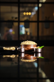 A richly detailed image featuring a pot filled with a savory rice dish, crowned with shredded cheese or garnishing. The pot is placed on a reflective surface, alongside a small metal bowl and green chili. The background is softly blurred, with warm lighting contributing to a cozy ambiance.