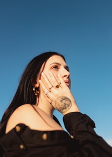 A woman is gazing at the sky with a contemplative expression. Her hand, adorned with rings, rests on her cheek, and a tattoo of the moon is visible on her hand. She is wearing a black off-the-shoulder top with button details, and large hoop earrings. The background is a clear blue sky, creating a peaceful and open atmosphere.