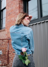A person with a short hairstyle stands outdoors against a backdrop of brick and metal siding. The individual wears a denim jacket and has makeup featuring red marks across their face. A pink rose is tucked into the waistband of their dark jeans.