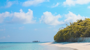 A serene beach scene with turquoise waters gently meeting the sandy shore. Lush green foliage covers the land on the right, while a traditional boat sails across the horizon under a clear blue sky scattered with fluffy clouds.