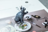 A ceramic figurine of a cat is positioned on top of a magazine or book page. The figurine is bluish-gray with a speckled pattern and portrays the cat in a sitting posture with one paw raised. It is placed on an image featuring a dining setting that includes a white plate with greens and a halved, peeled egg, a spoon, a pair of scissors, a bottle, and a smartphone on a dark surface.