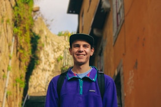A young person with a backpack is standing outdoors, smiling widely. They are wearing a purple pullover with turquoise accents and a dark cap. In the background, there's an outdoor stone staircase surrounded by rich greenery and orange-toned walls, suggesting a rustic urban setting.