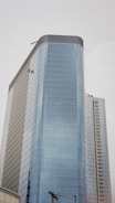A tall modern skyscraper with a sleek glass exterior, featuring multiple floors and a curved design. Two window cleaners are visible, dangling from their harnesses while performing maintenance on the building. The sky is overcast, creating a neutral background.