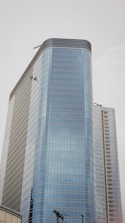 A tall modern skyscraper with a sleek glass exterior, featuring multiple floors and a curved design. Two window cleaners are visible, dangling from their harnesses while performing maintenance on the building. The sky is overcast, creating a neutral background.