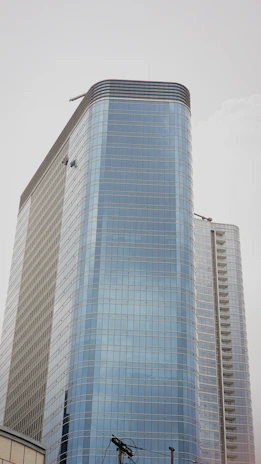 A tall modern skyscraper with a sleek glass exterior, featuring multiple floors and a curved design. Two window cleaners are visible, dangling from their harnesses while performing maintenance on the building. The sky is overcast, creating a neutral background.