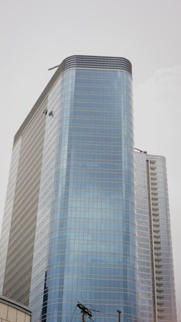 A tall modern skyscraper with a sleek glass exterior, featuring multiple floors and a curved design. Two window cleaners are visible, dangling from their harnesses while performing maintenance on the building. The sky is overcast, creating a neutral background.