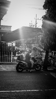 A person is riding a scooter with a large load, possibly carrying goods. The scene is set in an urban environment near a gated area with some bicycles in the background. A tree is on the right side, and the sun is creating a strong lens flare across the image.
