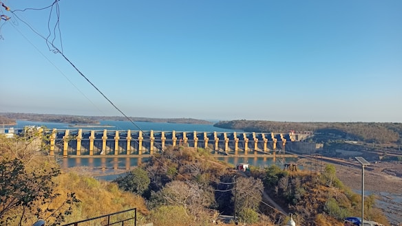 A vast dam stretches across a river with lush greenery on either side. The structure features multiple gates and is surrounded by a scenic landscape of water and hills.