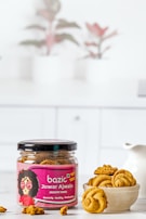 A jar labeled 'bazic Jowar Ajwain Jaggery Minis' sits on a white surface. The jar is open, revealing round golden-brown snacks inside. Beside the jar, a bowl holds several of these snacks. In the background, there are blurred images of potted plants, adding a decorative touch.