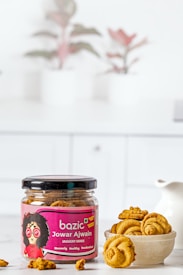 A jar labeled 'bazic Jowar Ajwain Jaggery Minis' sits on a white surface. The jar is open, revealing round golden-brown snacks inside. Beside the jar, a bowl holds several of these snacks. In the background, there are blurred images of potted plants, adding a decorative touch.