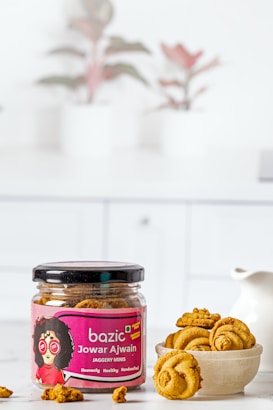 A jar labeled 'bazic Jowar Ajwain Jaggery Minis' sits on a white surface. The jar is open, revealing round golden-brown snacks inside. Beside the jar, a bowl holds several of these snacks. In the background, there are blurred images of potted plants, adding a decorative touch.