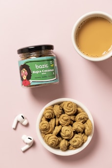 A jar of snack labeled 'Bazic Bajra Coconut Minis' is placed on a pink surface. Next to it is a small bowl filled with spiral-shaped cookies and a cup of tea with a light brown color. A pair of wireless earbuds are also placed nearby.