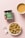 A jar of snack labeled 'Bazic Bajra Coconut Minis' is placed on a pink surface. Next to it is a small bowl filled with spiral-shaped cookies and a cup of tea with a light brown color. A pair of wireless earbuds are also placed nearby.