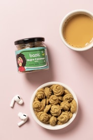 A jar of snack labeled 'Bazic Bajra Coconut Minis' is placed on a pink surface. Next to it is a small bowl filled with spiral-shaped cookies and a cup of tea with a light brown color. A pair of wireless earbuds are also placed nearby.