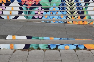 A series of stairs with vibrant, colorful floral patterns painted on the risers. The design features blue, pink, orange, and yellow flowers along with green foliage, creating a lively and artistic visual. The connected patterns extend across multiple steps with alternating colors.