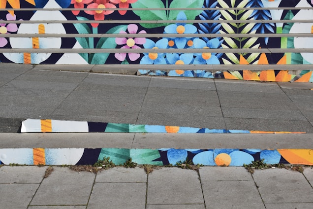 A series of stairs with vibrant, colorful floral patterns painted on the risers. The design features blue, pink, orange, and yellow flowers along with green foliage, creating a lively and artistic visual. The connected patterns extend across multiple steps with alternating colors.