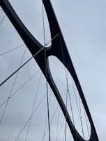A close-up view of a modern bridge's structural elements, featuring dark metal beams and tension cables set against a cloudy sky.