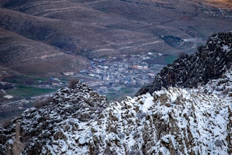 A snowy mountain landscape with rugged cliffs in the foreground and a small town nestled in a valley. The town is surrounded by rolling hills with patches of green, suggesting agricultural areas or grassy fields.