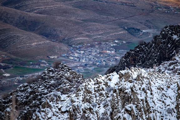 A snowy mountain landscape with rugged cliffs in the foreground and a small town nestled in a valley. The town is surrounded by rolling hills with patches of green, suggesting agricultural areas or grassy fields.
