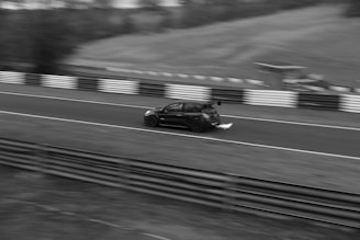A black car speeds along a race track, emitting flames from its exhaust. The surrounding environment includes a blurred background of grassy hills and a barrier along the track. The image captures a sense of motion and speed.