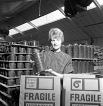 A person holding a patterned ceramic jug stands in a warehouse surrounded by stacks of similar jugs. Cardboard boxes labeled 'FRAGILE' and 'THIS SIDE UP' are in the foreground, indicating careful handling of the contents. The structure's high ceiling is visible, with skylights allowing natural light to illuminate the space.