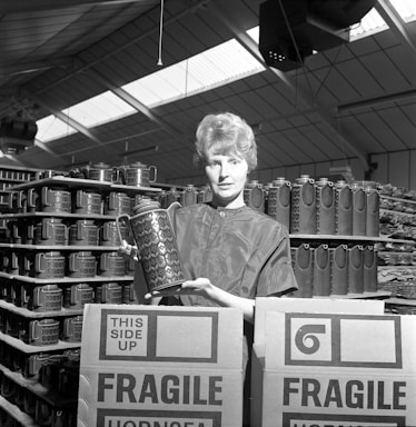 A person holding a patterned ceramic jug stands in a warehouse surrounded by stacks of similar jugs. Cardboard boxes labeled 'FRAGILE' and 'THIS SIDE UP' are in the foreground, indicating careful handling of the contents. The structure's high ceiling is visible, with skylights allowing natural light to illuminate the space.