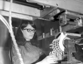 A person wearing glasses operates a piece of machinery that appears to be involved in manufacturing or decorating ceramics. The focus is on a mechanical device holding a large ceramic item with a distinctive geometric pattern. The environment looks industrial, with metal components and a dimly lit setting.