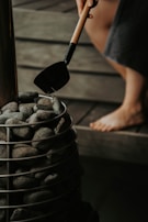 A person holding a ladle filled with water is pouring it over a pile of smooth gray stones placed in a metal basket. The scene suggests a spa or sauna environment, with wooden surfaces visible in the background and a barefoot individual's leg partially shown.