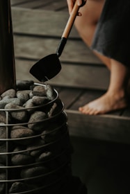 A person holding a ladle filled with water is pouring it over a pile of smooth gray stones placed in a metal basket. The scene suggests a spa or sauna environment, with wooden surfaces visible in the background and a barefoot individual's leg partially shown.