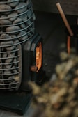A compact wood-burning stove with a visible flame inside, surrounded by a metal basket filled with smooth gray stones. There is a wooden stick resting inside a metal container nearby.