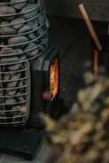 A compact wood-burning stove with a visible flame inside, surrounded by a metal basket filled with smooth gray stones. There is a wooden stick resting inside a metal container nearby.
