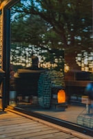 A warm, cozy interior is visible through a large glass window, reflecting the outdoors. Inside, a modern wood-burning stove with a stockpile of neatly arranged logs emits a soft glow, contributing to an inviting ambiance. The reflection of trees and a person can be seen, suggesting a connection between nature and the comfortable indoor setting.