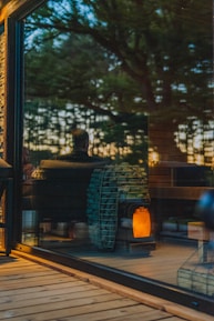 A warm, cozy interior is visible through a large glass window, reflecting the outdoors. Inside, a modern wood-burning stove with a stockpile of neatly arranged logs emits a soft glow, contributing to an inviting ambiance. The reflection of trees and a person can be seen, suggesting a connection between nature and the comfortable indoor setting.