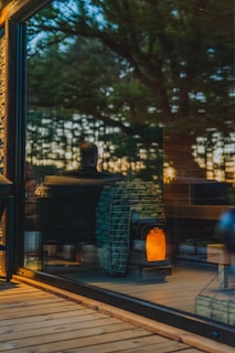 A warm, cozy interior is visible through a large glass window, reflecting the outdoors. Inside, a modern wood-burning stove with a stockpile of neatly arranged logs emits a soft glow, contributing to an inviting ambiance. The reflection of trees and a person can be seen, suggesting a connection between nature and the comfortable indoor setting.