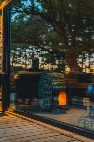 A warm, cozy interior is visible through a large glass window, reflecting the outdoors. Inside, a modern wood-burning stove with a stockpile of neatly arranged logs emits a soft glow, contributing to an inviting ambiance. The reflection of trees and a person can be seen, suggesting a connection between nature and the comfortable indoor setting.