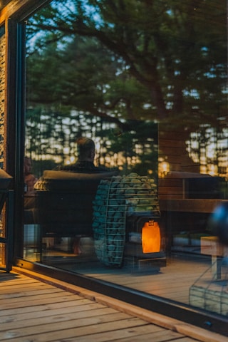 A warm, cozy interior is visible through a large glass window, reflecting the outdoors. Inside, a modern wood-burning stove with a stockpile of neatly arranged logs emits a soft glow, contributing to an inviting ambiance. The reflection of trees and a person can be seen, suggesting a connection between nature and the comfortable indoor setting.