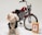 A small, fluffy dog wearing a black leather jacket stands confidently beside a retro-style motorcycle with a bold red gas tank and chrome details. In front of the motorcycle is a packet labeled 'Dad Grass'. The background is plain, enhancing the focus on the dog, motorcycle, and packaging.