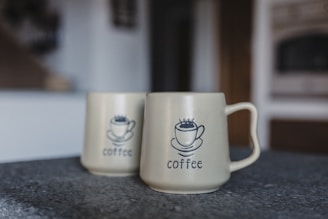 Two ceramic mugs with a simple coffee cup illustration and the word 'coffee' printed underneath. They are placed on a textured surface in a softly lit room, with a blurred background suggesting an indoor setting.