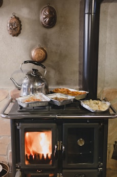 A traditional wood-burning stove with a visible fire inside the stove door. Several aluminum trays with cooked food and a shiny metal kettle are placed on top of the stove. The background features a textured wall adorned with vintage, decorative copper molds.