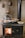 A traditional wood-burning stove with a visible fire inside the stove door. Several aluminum trays with cooked food and a shiny metal kettle are placed on top of the stove. The background features a textured wall adorned with vintage, decorative copper molds.