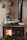 A traditional wood-burning stove with a visible fire inside the stove door. Several aluminum trays with cooked food and a shiny metal kettle are placed on top of the stove. The background features a textured wall adorned with vintage, decorative copper molds.