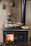 A traditional wood-burning stove with a visible fire inside the stove door. Several aluminum trays with cooked food and a shiny metal kettle are placed on top of the stove. The background features a textured wall adorned with vintage, decorative copper molds.